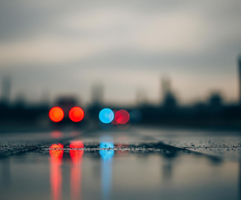 Rozmyta fotografia przedstawia mokrą asfaltową nawierzchnię ulicy lub drogi, na której odbijają się światła samochodów. Na pierwszym planie widać ciemny, ziarnisty asfalt z drobnymi kroplami wody, tworzącymi wyraźną linię horyzontu. Poniżej tej linii, w odbiciu na mokrej powierzchni, widoczne są intensywne smugi światła: dwa czerwone po lewej i jeden niebieski po prawej. Powyżej linii horyzontu, w tle, widoczne są rozmyte światła samochodów – dwa czerwone po lewej i dwa niebieskie po prawej – oraz niewyraźne, ciemne sylwetki budynków lub drzew. Całość utrzymana jest w chłodnej tonacji szarości, błękitów i czerwieni, z dominującym efektem bokeh dla świateł w tle.