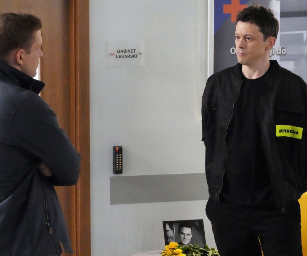 Daniel (Marcin Bikowski) jako ochroniarz, w czarnej kurtce z żółtym napisem OCHRONA na ramieniu, rozmawia z Hubertem (Marek Molak) w ciemnej kurtce. Scena ma miejsce w szpitalu, obok drzwi z napisem GABINET LEKARSKI, na tle plakatu z lekarką. O dramatycznych losach bohaterów przeczytasz na Super Seriale.
