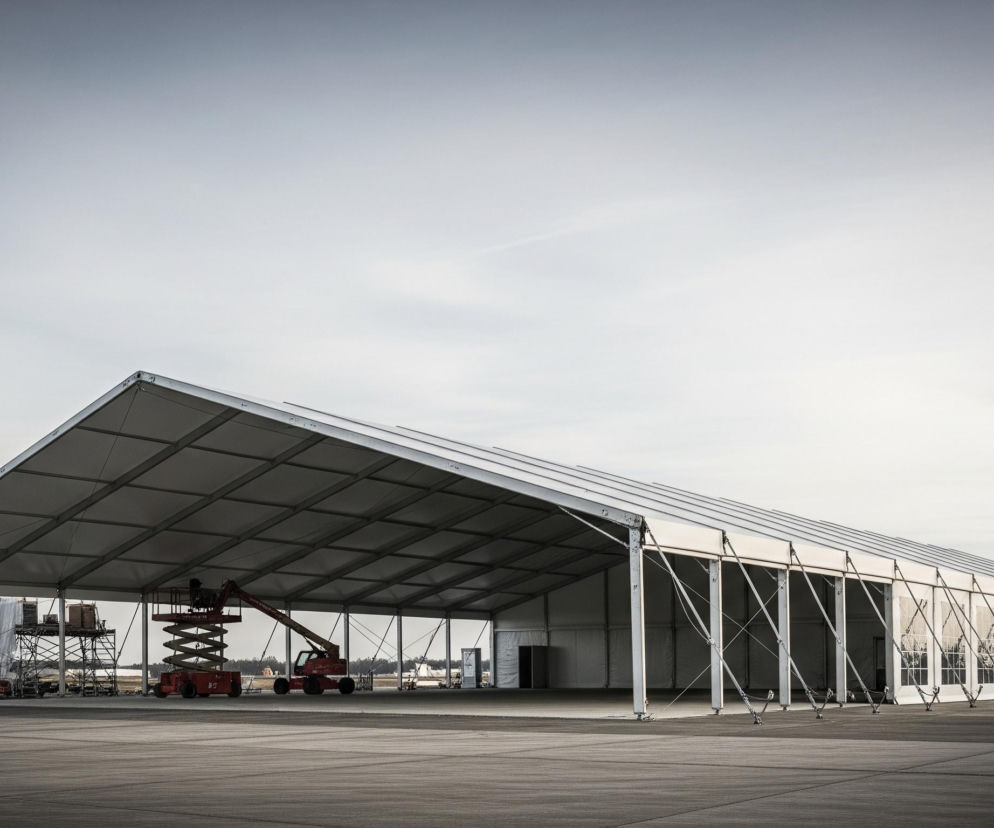 Duży, długi namiot w jasnych kolorach, prawdopodobnie biały lub szary, z dachem dwuspadowym i otwartą boczną ścianą, dominuje środek obrazu. Wewnątrz namiotu, po lewej stronie, widoczne są dwie pomarańczowe lub czerwone maszyny podnośnikowe, jedna typu nożycowego, a druga teleskopowego, a także struktury rusztowania. Po prawej stronie namiotu, na zewnątrz, znajduje się kolejna pomarańczowa maszyna podnośnikowa, a cała konstrukcja stoi na obszernej, szarej, asfaltowej powierzchni. Niebo nad namiotem jest jasnoszare i bezchmurne.