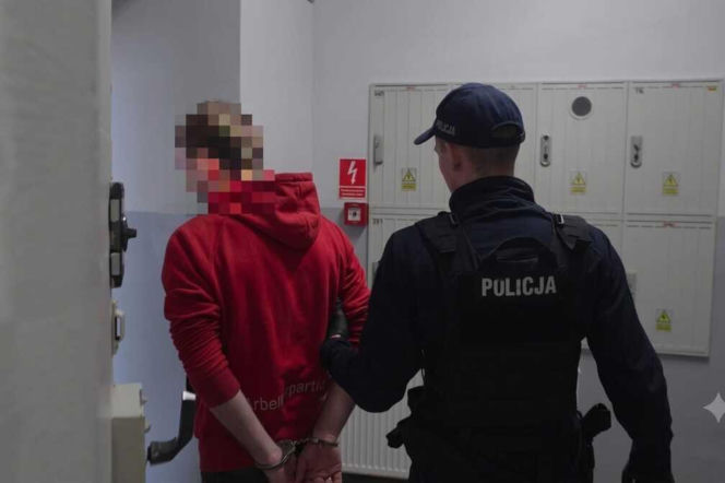 Policjant prowadzi skutego kajdankami 23-latka w czerwonej bluzie. O aresztowaniu sprawcy wypadku przeczytasz na SE.