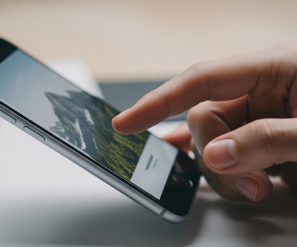 Dłoń trzyma smartfon z aluminiową obudową, a kciuk dotyka ekranu. Na ekranie wyświetlany jest krajobraz górski z zielonymi zboczami i szarymi szczytami pod niebieskim niebem. W tle widoczny jest rozmyty, jasny blat.