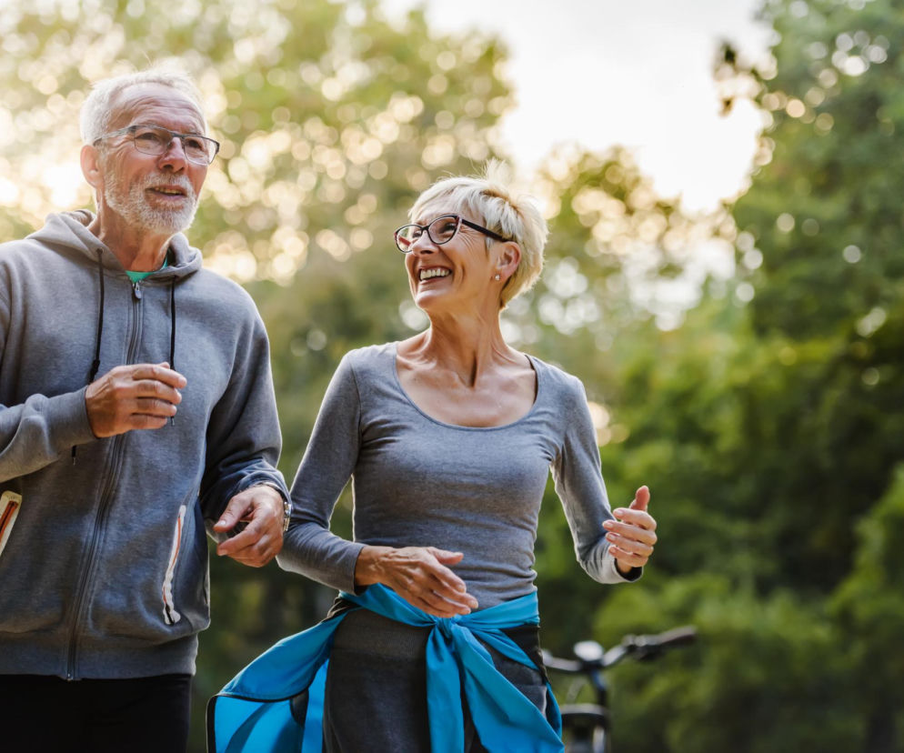 Para dojrzałych osób w sportowych ubraniach biegnie parkową alejką. Kobieta w okularach ma rozpromienioną twarz i patrzy w bok, a mężczyzna obok niej skupiony na biegu. Ich aktywność fizyczna po diagnozie raka może wydłużyć życie, o czym przeczytasz na Poradnik Zdrowie.
