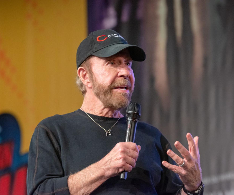 Aktor Chuck Norris w czarnej czapce z daszkiem i bluzie, z brodą, trzymający mikrofon i gestykulujący. Zdjęcie ilustruje temat zdrowia i nagłych incydentów medycznych, o których można przeczytać więcej na portalu Poradnik Zdrowie.