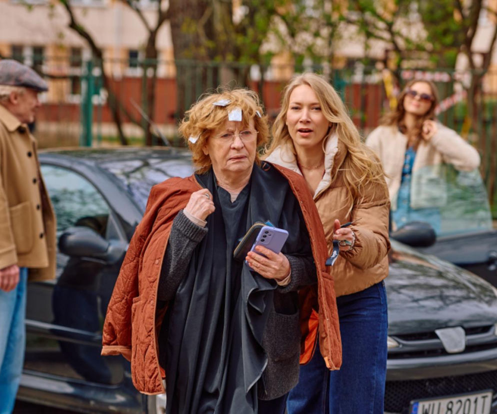 Krystyna (Małgorzata Knothe) z córką Łucją (Magdalena Kaczmarek) po wyjściu od fryzjera. Krystyna ma na głowie papierki do utrwalenia fryzury. W tle widać Juliusza (Daniel Olbrychski) i Andżelę (Joanna Opozda) w samochodzie. Zobacz streszczenia odcinków serialu Na Wspólnej na portalu Super Seriale.