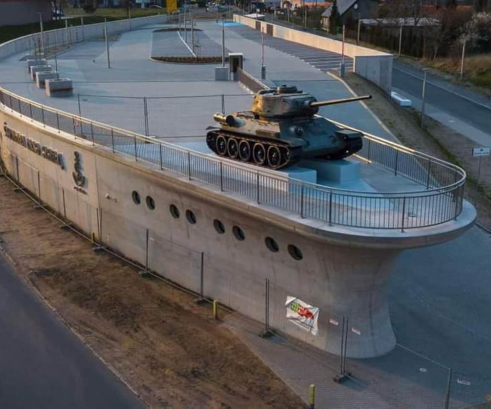 Nietypowy Park&Ride w Ścinawie, betonowy budynek przypominający statek z czołgiem T-34 na dziobie. Obiekt otoczony pustymi miejscami parkingowymi, w tle widoczne fragmenty miejskiej zabudowy. Więcej o wyzwaniach architektury przeczytasz na Architektura Murator Plus.