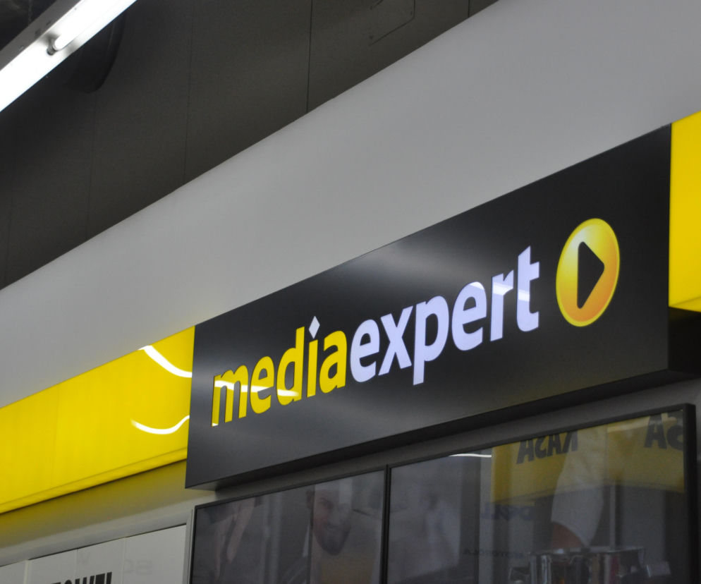 Podświetlany, czarno-żółty szyld z logo Media Expert, widoczny wewnątrz sklepu, symbolizujący otwarte drzwi dla klientów. Artykuł na Super Biznes informuje o godzinach otwarcia sklepów RTV AGD w długi weekend czerwcowy 2026.