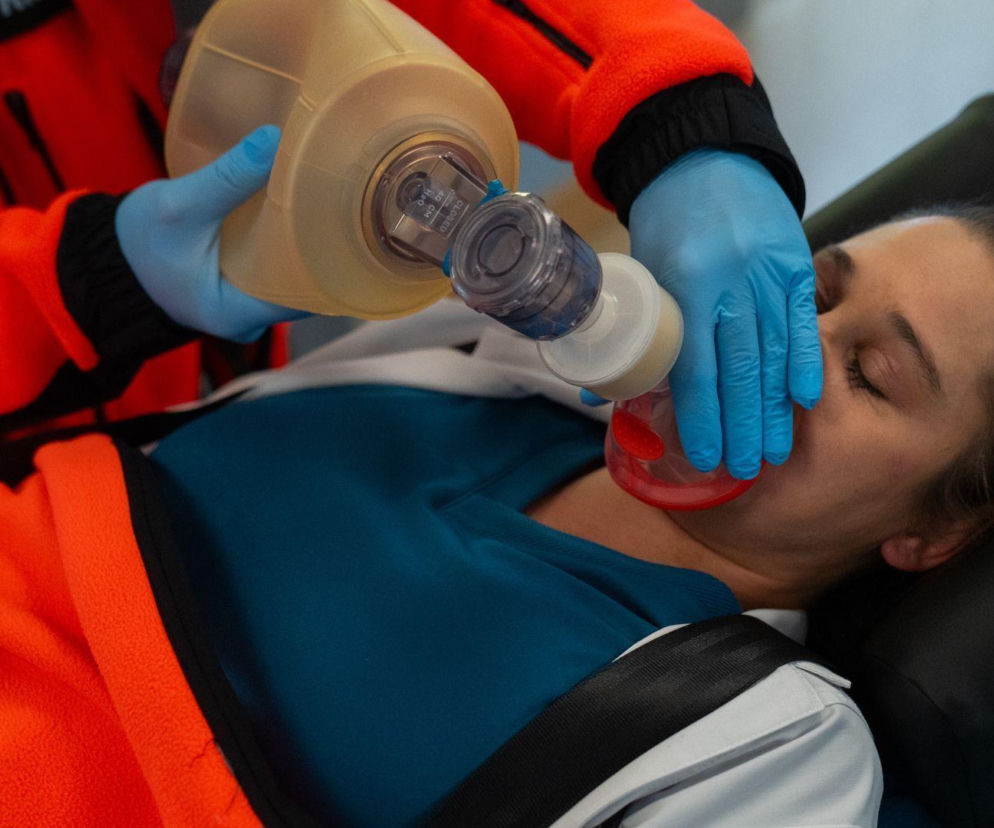 Ratownik medyczny w pomarańczowym kombinezonie ratuje życie kobiecie z brązowymi włosami, trzymając maskę tlenową na jej twarzy. Kobieta leży na noszach, ubrana w niebieską bluzkę. Scena z serialu Na dobre i na złe, więcej o tym na Super Seriale.