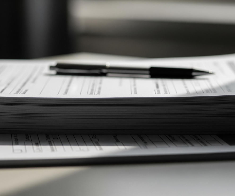 Na pierwszym planie, w centrum kadru, leży stos białych kartek papieru z wydrukowanym tekstem i liniami, tworzącymi formularze lub dokumenty. Na wierzchu tego stosu umieszczony jest czarny długopis o metalicznym połysku, skierowany końcówką w prawą stronę. Stos kartek rzuca cień na powierzchnię stołu lub biurka, na którym spoczywa, a tło za nim jest ciemne i rozmyte, co sugeruje głębię ostrości skupioną na dokumentach i długopisie.