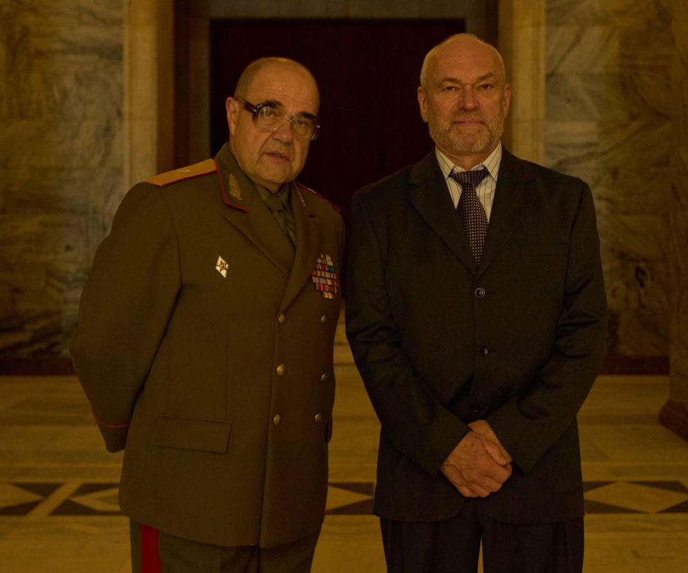 Cezary Żak jako generał Dubynin i Mirosław Haniszewski jako Sadowski w serialu Wojna zastępcza. Obaj mężczyźni w eleganckich strojach, stoją na tle marmurowych ścian i podłogi. Więcej o obsadzie i bohaterach przeczytasz na Super Seriale.