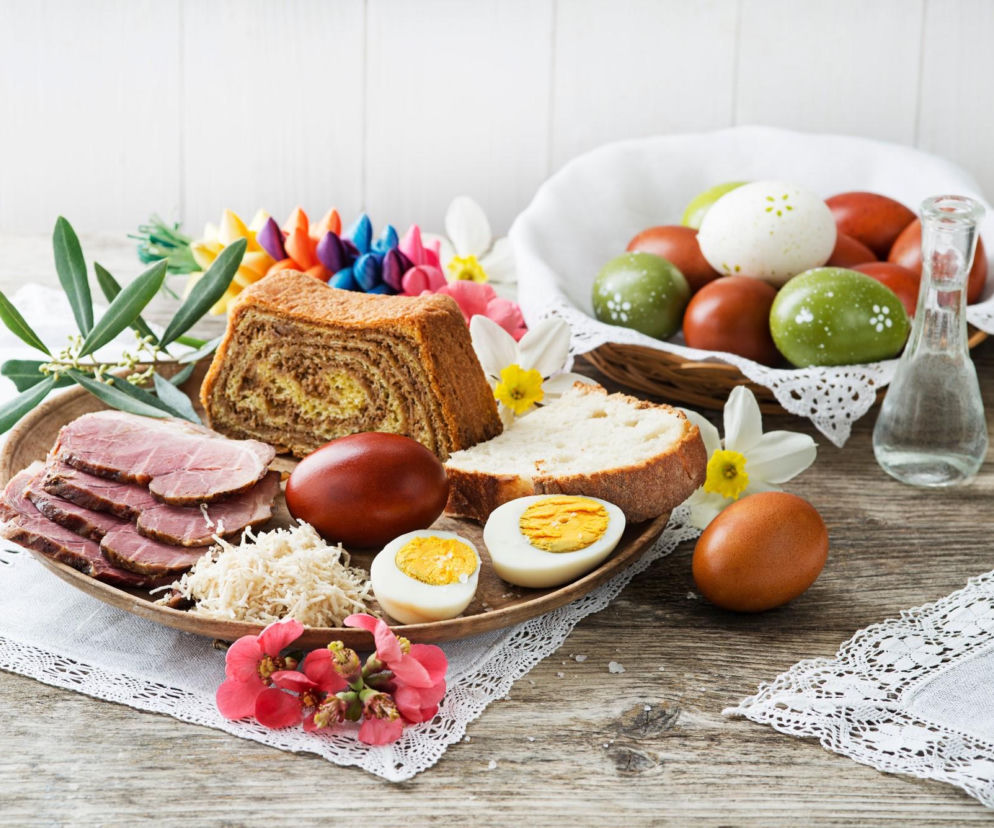Wielkanocny stół z potrawami, takimi jak szynka, jajka, chleb i babka, otoczony kolorowymi pisankami i wiosennymi kwiatami, symbolizujący świąteczne wydatki i porównanie cen w sklepach na Super Biznes.