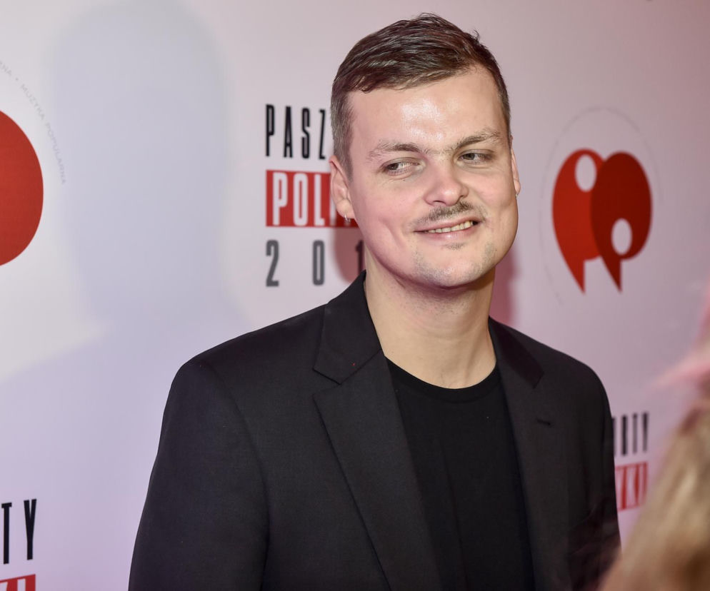 Błażej Król w ciemnej marynarce uśmiecha się na ściance wydarzenia Paszporty Polityki. O artyście w Azja Express przeczytasz w Eska.