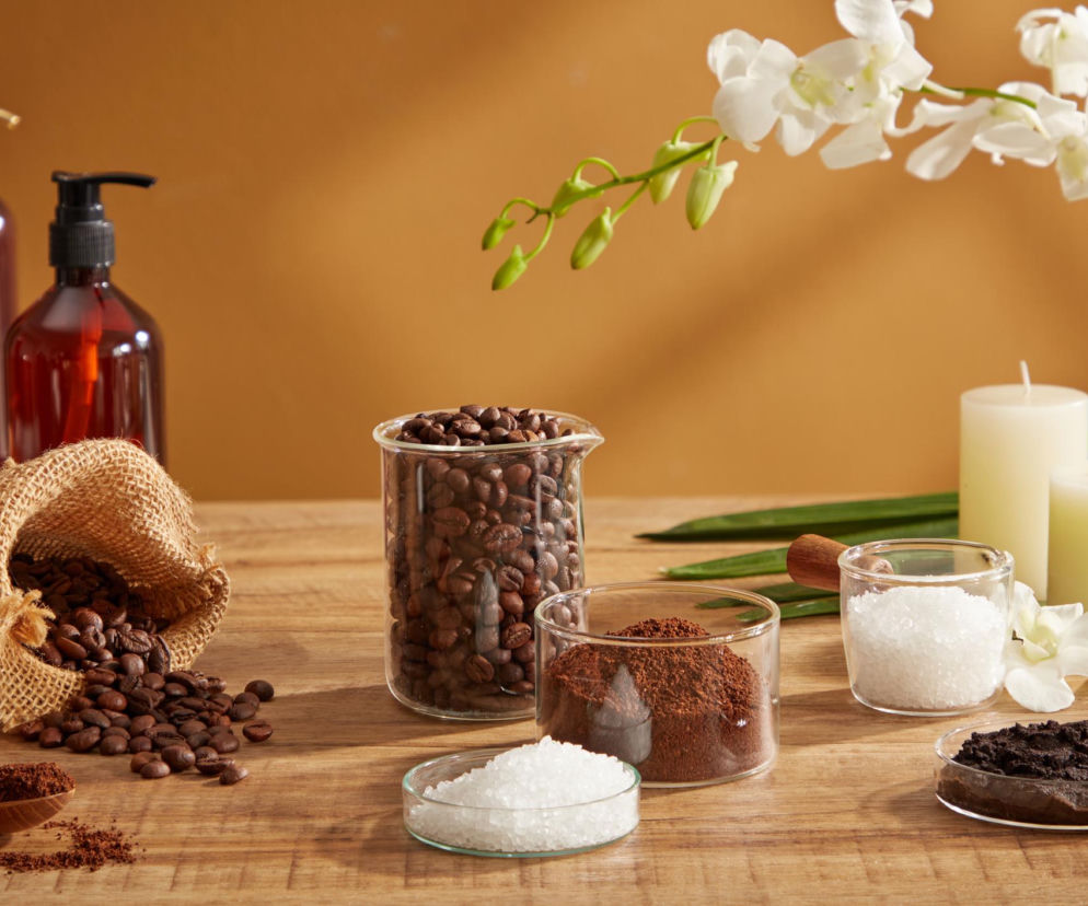 Peelingi kawowe, cukrowe i solne na drewnianym stole: rozsypane ziarna kawy z worka, naczynia z kawą mieloną, solą i gotowym peelingiem, obok świece i orchidea. Przeczytaj, jak wybrać najlepszy peeling na naszym portalu.