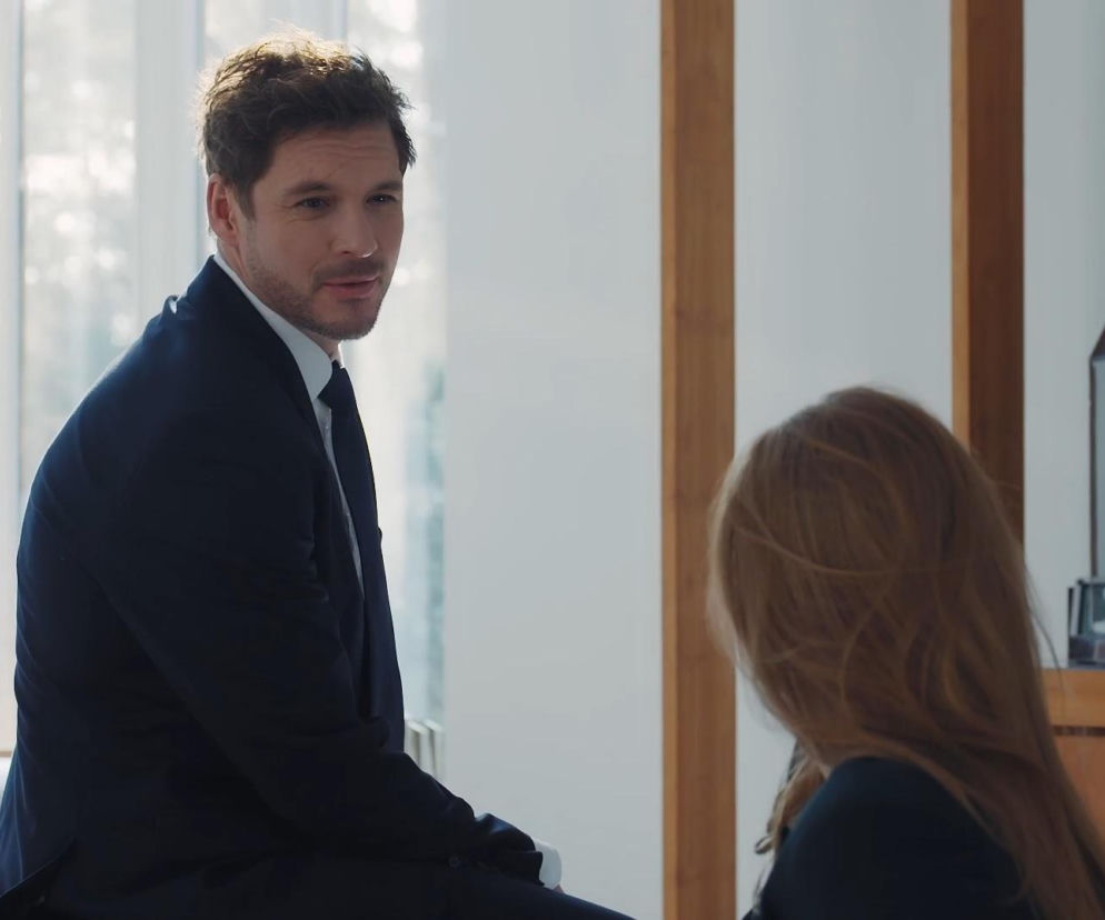 Mikołaj Roznerski jako Marcin Chodakowski w granatowym garniturze z krawatem, siedzący tyłem do okna, spogląda w prawo na Elżbietę Domańską (Katarzyna Dąbrowska) z długimi blond włosami, której widać tylko tył głowy. Scena z serialu M jak miłość na Super Seriale.