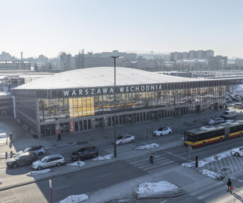 Budynek Dworca Wschodniego w Warszawie z szeroką fasadą, otoczony ulicą z autami i autobusami oraz postojem taksówek. W tle widać fragment Stadionu Narodowego. Więcej o planach połączenia dworca z 3. linią metra przeczytasz na Architektura Murator Plus.