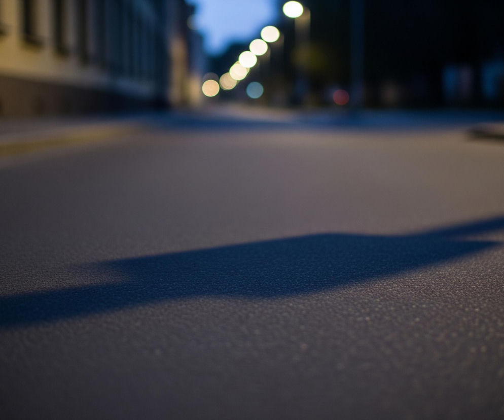 Ulica o zmierzchu lub nocą jest głównym elementem obrazu, widziana z niskiej perspektywy. Szara, ziarnista powierzchnia asfaltu z ciemnoniebieskim cieniem przebiegającym ukośnie przez środek zajmuje większą część kadru. Po lewej stronie widać niewyraźne, żółtawe fasady budynków, a w oddali, wzdłuż zakręcającej drogi, rozciąga się rząd rozmytych, okrągłych świateł ulicznych w odcieniach żółci i bieli.