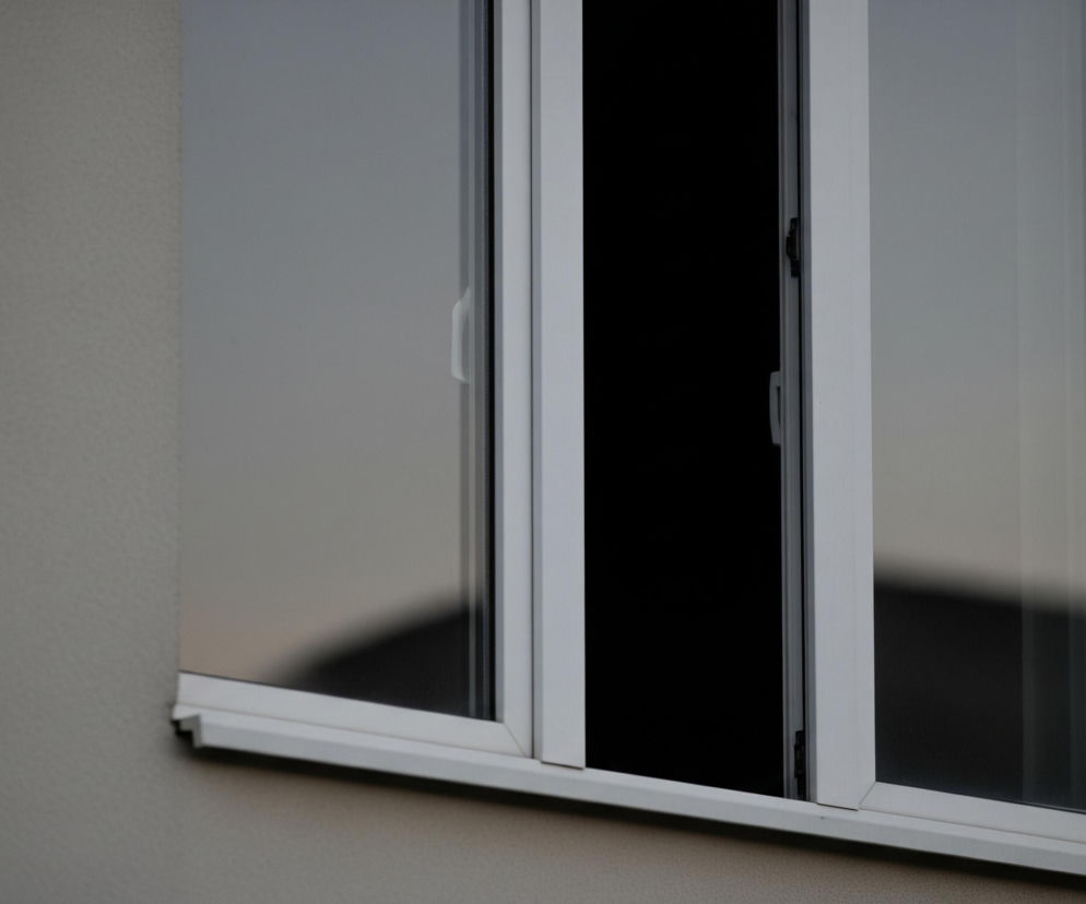 Fragment jasnoszarej, teksturowanej ściany zajmuje lewą część kadru, płynnie przechodząc w białe ramy okienne. Okno składa się z dwóch prostokątnych skrzydeł, z czego lewe jest zamknięte, a prawe uchylone w kierunku wnętrza, tworząc ciemną, pionową szczelinę. Szyby, odbijając otoczenie, prezentują odcienie szarości i czerń, z subtelnymi rozmyciami odzwierciedlającymi elementy z zewnątrz.