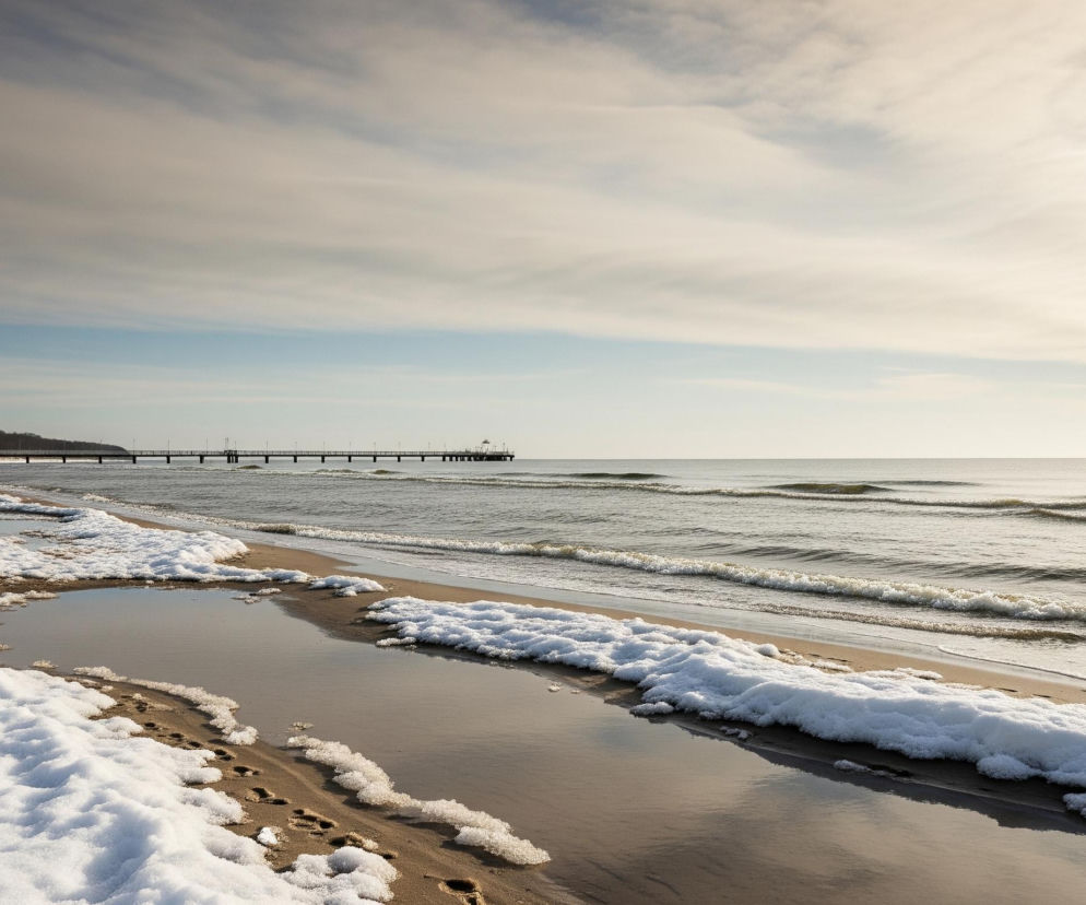 Szeroki kadr przedstawia zimową plażę z pomostem rozciągającym się w dal, pod częściowo zachmurzonym niebem. Na pierwszym planie, po lewej stronie, brązowy piasek pokryty jest nieregularnymi płatami białego śniegu i wodą, w której odbija się jasne niebo. Widać także ślady stóp na śniegu i mokrym piasku. Dalej, po prawej, ciemnoszare morze ze spokojnymi, falującymi falami styka się z piaszczystym brzegiem, a w tle widać długi, drewniany pomost wsparty na palach, prowadzący w głąb morza. Linię brzegową po lewej stronie obrazu zamyka pas nagich drzew.