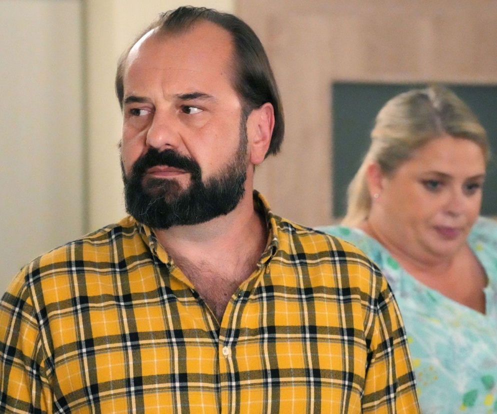 Borys (Jakub Wieczorek) z zamyślonym wyrazem twarzy, ubrany w żółto-czarną koszulę w kratę, obok Aldona (Elżbieta Romanowska) w tle, patrząca w dół. Scena z odcinka serialu Barwy szczęścia, o którym można przeczytać więcej na portalu Super Seriale.