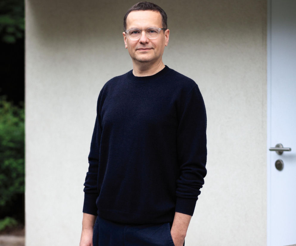 Architekt Rafał Mazur w okularach i ciemnym swetrze stoi przed budynkiem. O pracy architekta przeczytasz na Architektura.