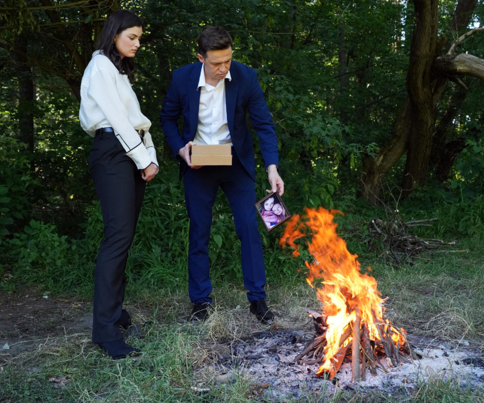 Piotr Ligienza jako Błażej i Maria Świłpa jako Weronika, stojący w lesie obok płonącego ogniska. Błażej trzyma pudełko z kartonu oraz ramkę ze zdjęciem, które wrzuci do ognia. Weronika stoi obok niego. Dramatyczny gest to symboliczne zerwanie z przeszłością, o czym przeczytasz na stronie Super Seriale.