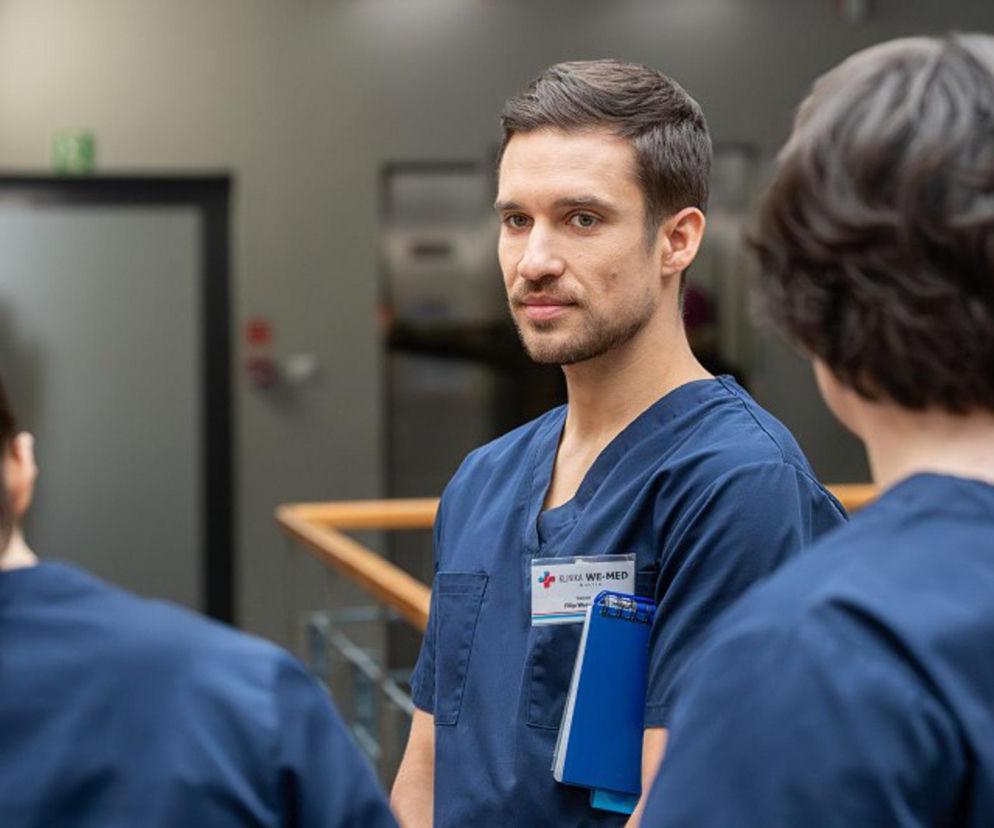 Lekarz Filip w niebieskim uniformie medycznym z identyfikatorem Klinika We-Med, trzymający niebieską podkładkę z kartkami, stoi w otoczeniu innych pracowników szpitala. Scena nawiązuje do odcinka Pierwszej miłości o masowym zatruciu, o którym przeczytasz na Super Seriale.