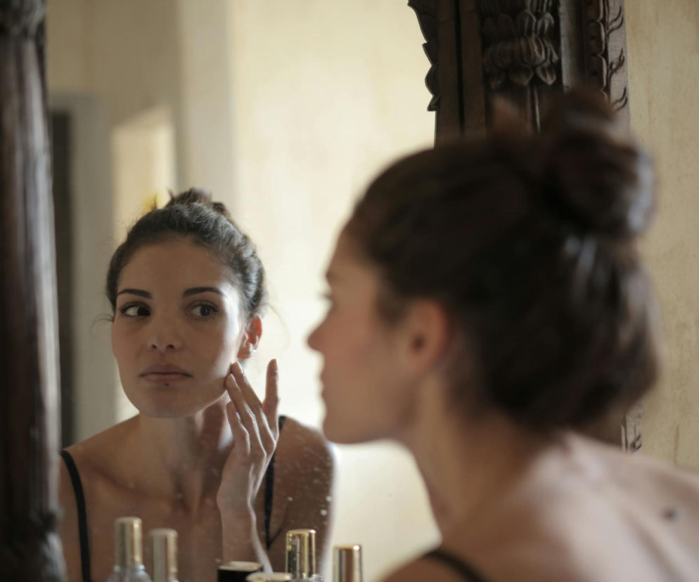 Kobieta z upiętymi włosami w lustrze, delikatnie dotykająca skóry na policzku, z zamyśleniem oceniająca stan swojej cery. Odzwierciedla troskę o wygląd skóry twarzy. Na Poradnik Zdrowie znajdziesz wskazówki, jak dbać o skórę i unikać krostek.