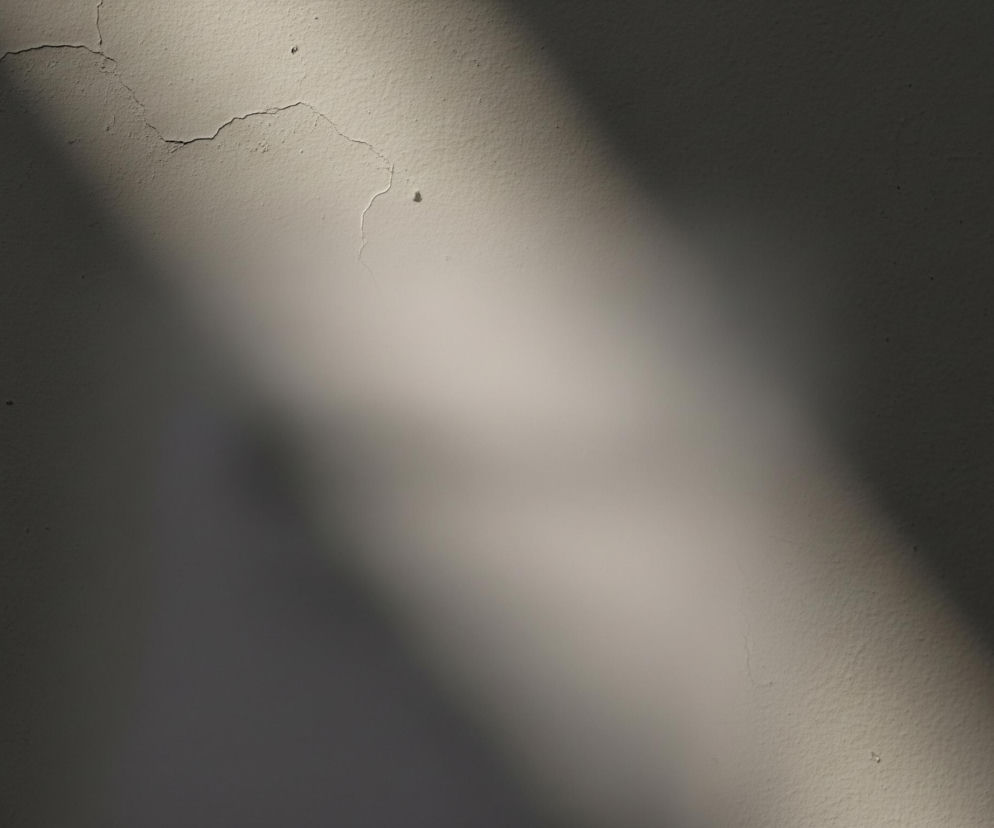 Tekstura ściany, pokrytej jasną farbą o beżowym odcieniu, jest nierówna i lekko chropowata, z widocznymi drobnymi pęknięciami i zaciekami. Przekątny pas światła przechodzi przez środek kompozycji, tworząc wyraźny kontrast między intensywnie oświetlonym obszarem a zacienionymi fragmentami. W górnej lewej części obrazu widoczne są pęknięcia biegnące poziomo i w dół, natomiast dolna prawa część charakteryzuje się delikatniejszą teksturą bez wyraźnych uszkodzeń.