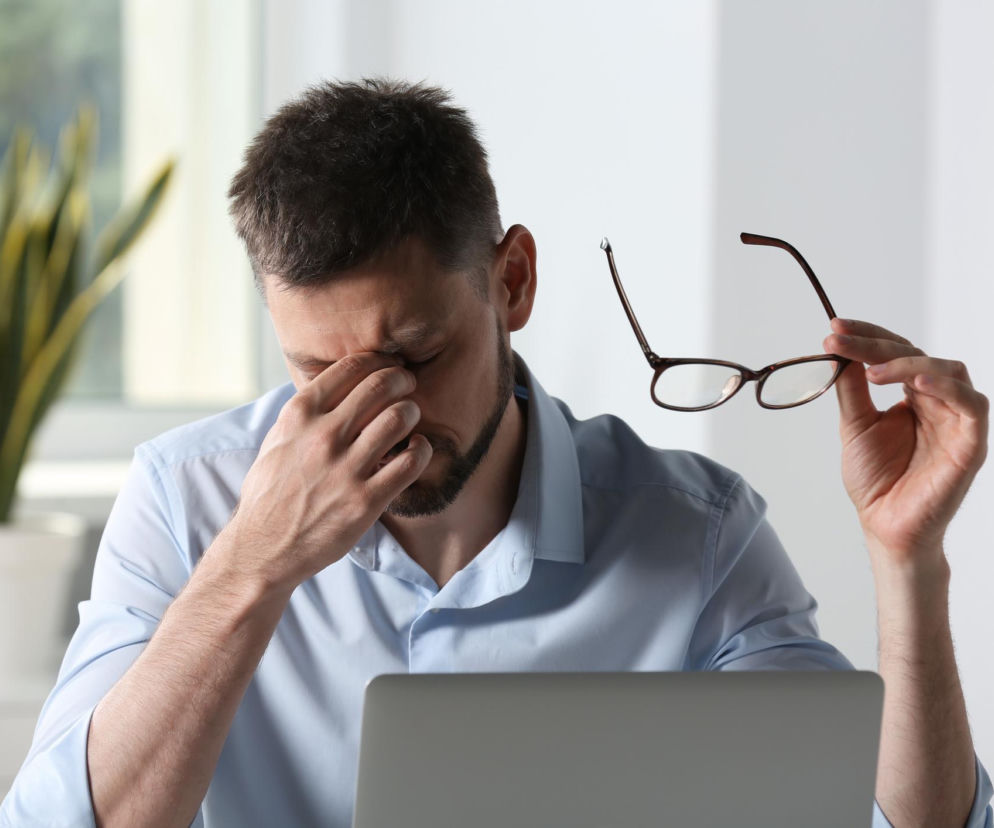 Mężczyzna w jasnej koszuli masuje skronie, trzymając okulary w drugiej dłoni, siedzi przed laptopem. Przedstawia zmęczenie wzroku po pracy przy komputerze, wskazując na potrzebę ulgi, o której można przeczytać na Poradnik Zdrowie.