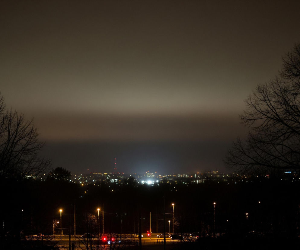 Nocny krajobraz miasta, z centralnie umieszczonym pasmem oświetlonych budynków na horyzoncie, rozświetla ciemne niebo, które stopniowo jaśnieje ku dołowi. Na pierwszym planie, po lewej i prawej stronie, dominują sylwetki nagich drzew z rozgałęzionymi konarami, tworząc ciemną ramę dla widoku. Poniżej miasta, w tle, widoczne są pojedyncze, żółte światła latarni ulicznych oraz czerwone światła drogowe, sugerujące ruch uliczny w oddali.