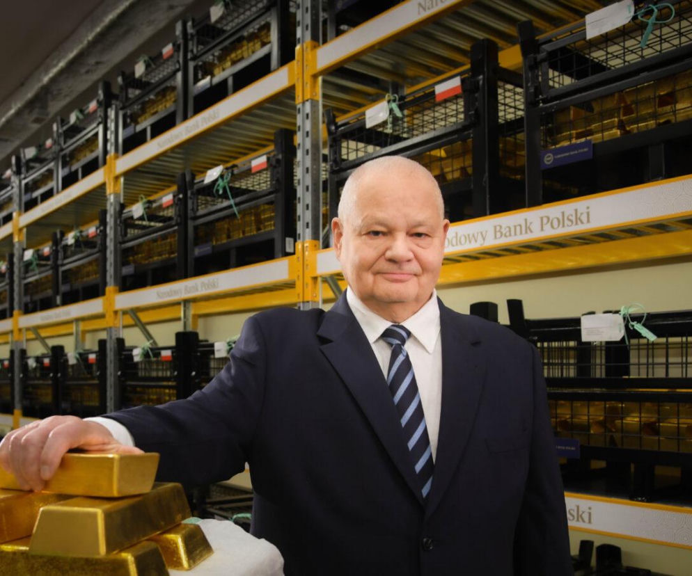 Prezes NBP Adam Glapiński w skarbcu banku, opierający rękę na stosie złotych sztabek. W tle regały pełne złota z flagami Polski, z opisem Narodowy Bank Polski. Artykuł o celach rezerw kruszcu przeczytasz na Super Biznes.