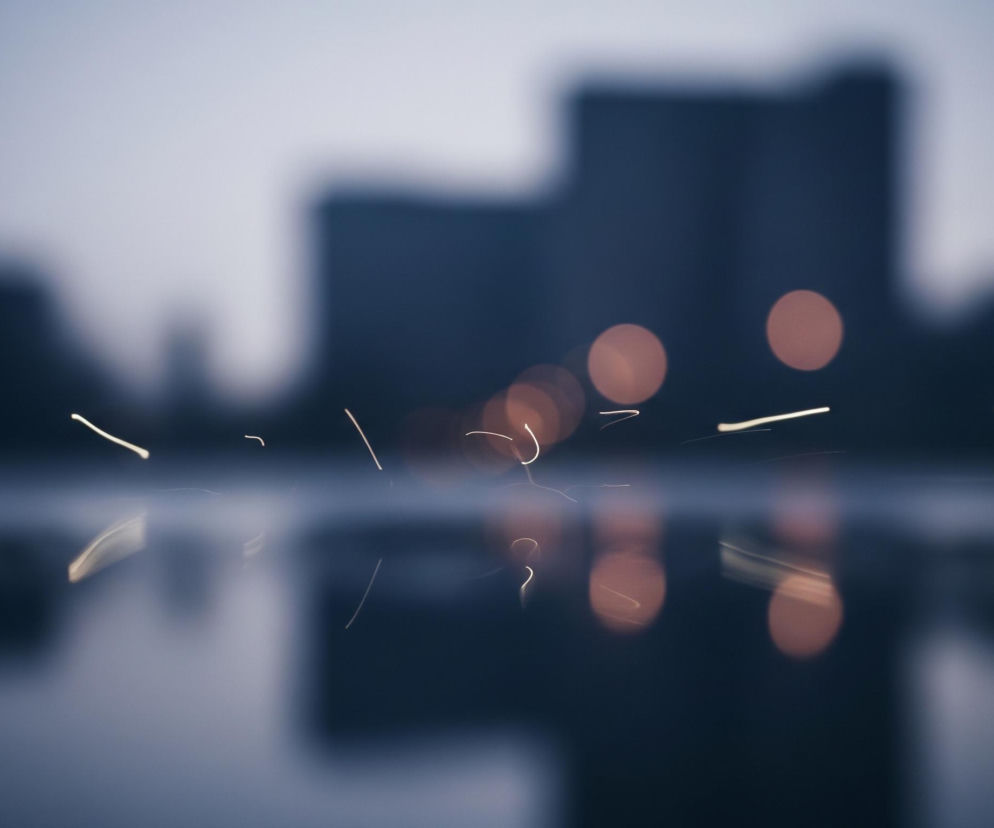 Na pierwszym planie widoczne są rozmyte, poziome, białe smugi światła o różnej długości, odbijające się w lekko ciemniejszej tafli pod spodem. Za nimi, w centrum obrazu, rozświetlona jest seria okrągłych, rozmytych punktów światła w kolorze pomarańczowym, które również mają swoje odbicia. W tle znajdują się ciemne, niewyraźne sylwetki budynków, przypominające zarys miejskiej panoramy, a całe otoczenie utrzymane jest w chłodnych odcieniach szarości i błękitu.