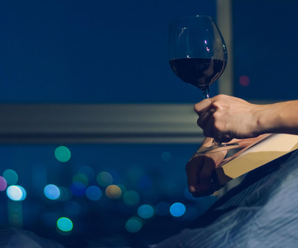 Dłoń trzymająca kieliszek czerwonego wina w wieczornym otoczeniu, na tle rozmytych świateł miasta. Ilustracja odzwierciedla temat wpływu wieczornego drinka na sen i poranne zmęczenie, o czym przeczytasz więcej na Poradnik Zdrowie.