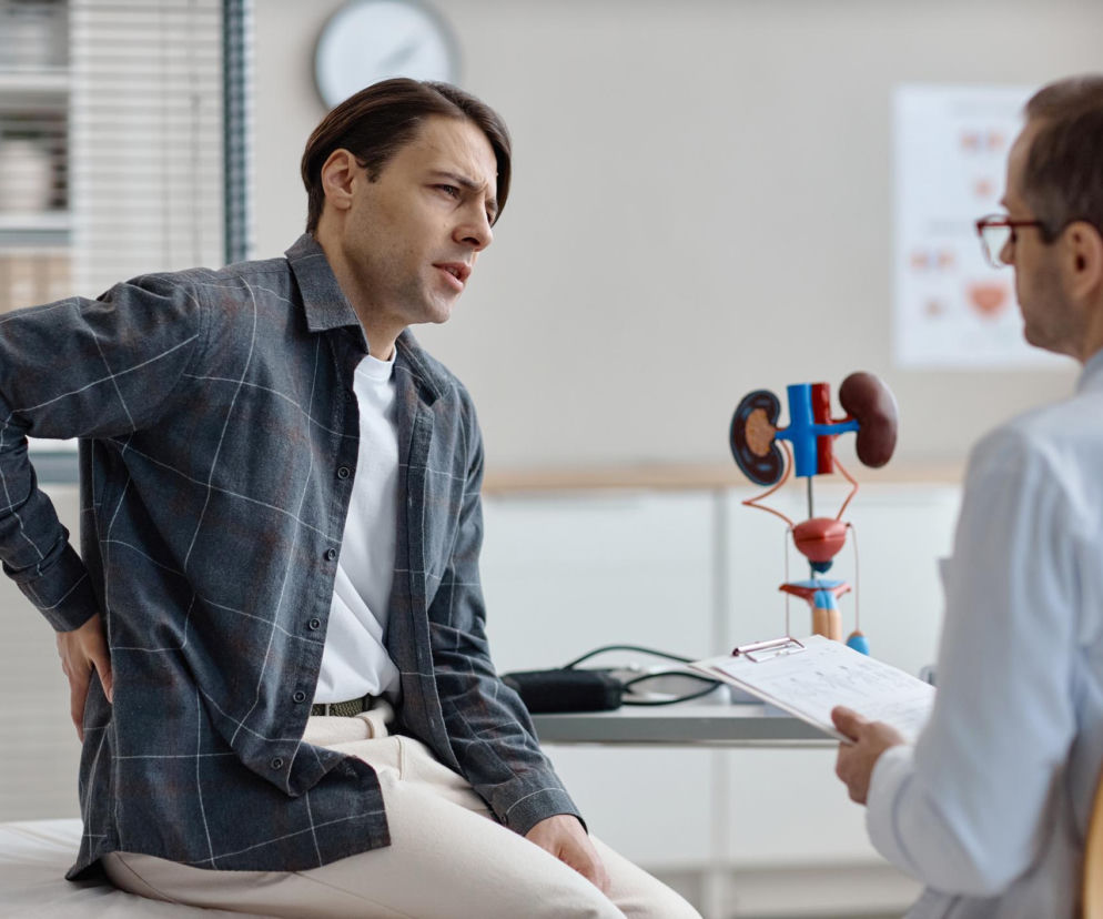 Mężczyzna z bólem pleców, siedzący podczas wizyty u lekarza w gabinecie urologicznym, obok model układu moczowego. Ilustracja objawów wodonercza i potrzeby szybkiej diagnostyki, o której przeczytasz na portalu Poradnik Zdrowie.