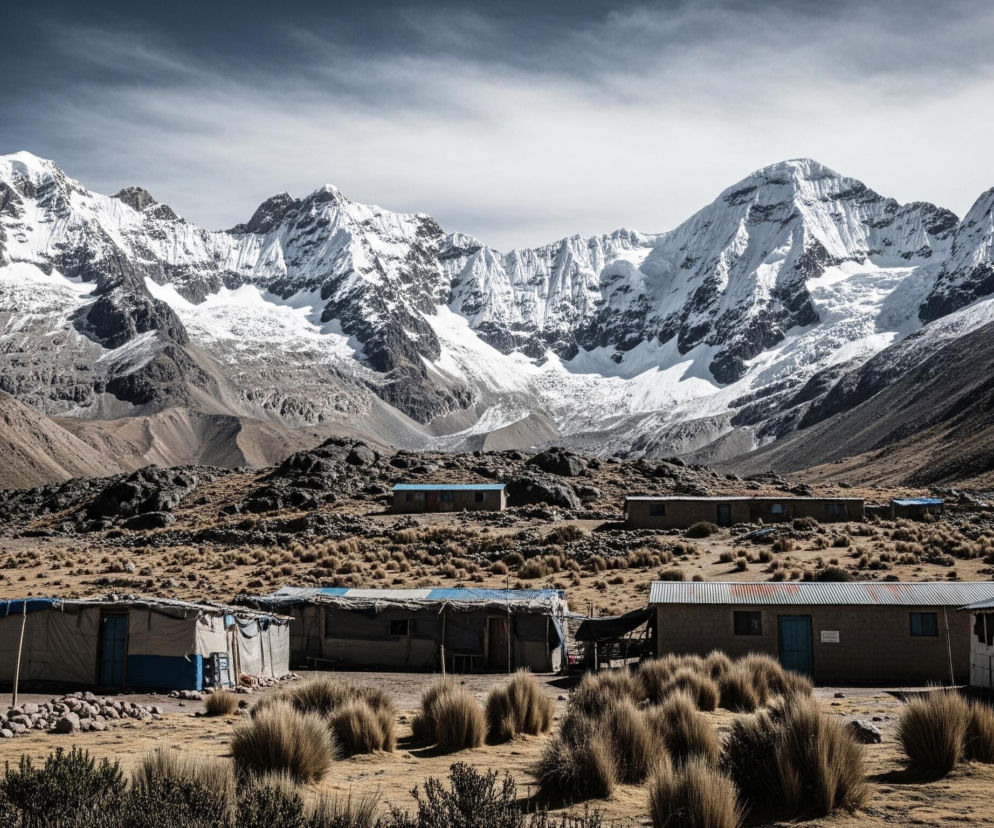 Na pierwszym planie rozległe, suche, pagórkowate tereny pokryte kępami wyschniętej trawy i rozsianymi skałami. Widać szereg prostych, niskich budynków z szarych lub jasnobrązowych materiałów, niektóre z niebieskimi drzwiami lub fragmentami dachów pokrytymi niebieską plandeką, tworzących niewielką osadę. W tle dominują potężne, skaliste góry, których szczyty i zbocza są częściowo pokryte śniegiem i lodowcami, widoczne pod zachmurzonym niebem.
