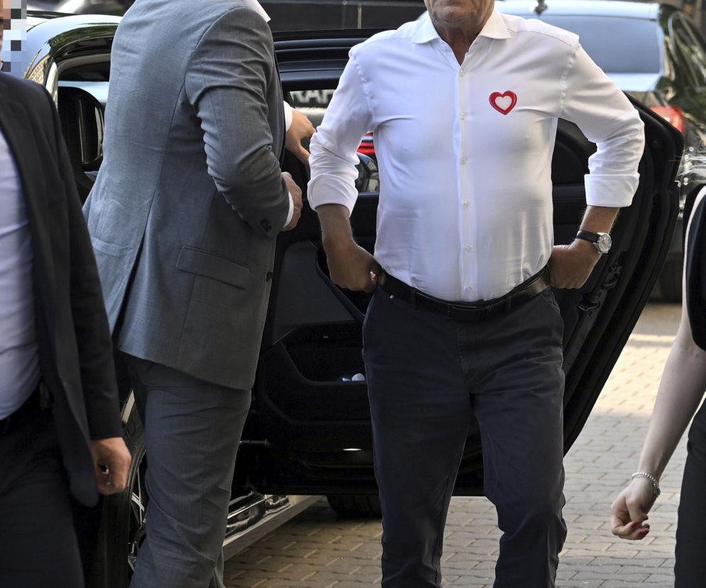 Premier Donald Tusk w białej koszuli z czerwonym sercem stoi przy samochodzie. O cenach paliw przeczytasz na Eska.