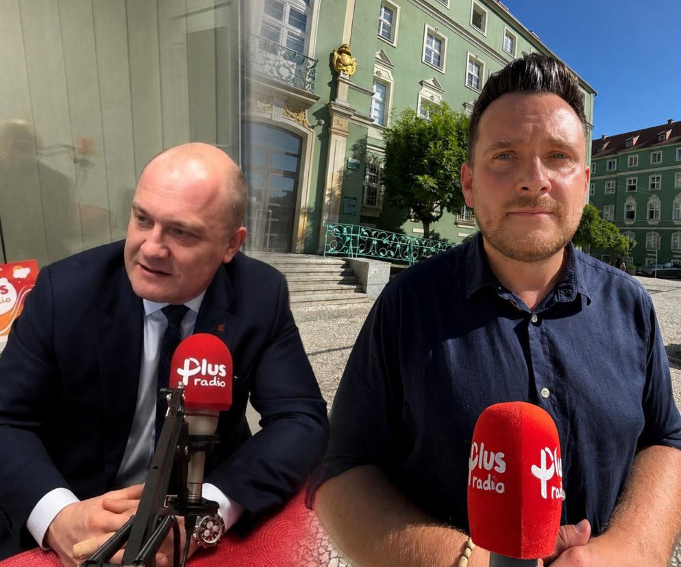 Prezydent Piotr Krzystek w garniturze rozmawia do mikrofonu Radia Plus, obok Radny Krzysztof Romianowski z mikrofonem tego samego radia na tle zielonego budynku. Artykuł o konflikcie w samorządzie przeczytasz na naszym portalu.