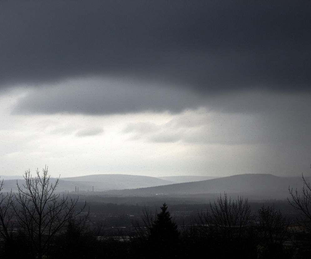 Horyzontalna fotografia przedstawia zimowy, pochmurny krajobraz z ciemnym, dominującym niebem w górnej części kadru, przechodzącym w jaśniejsze odcienie szarości w kierunku linii horyzontu. Na pierwszym planie, u dołu obrazu, widoczne są czarne, nagie konary drzew bez liści, częściowo zasłaniające widok na rozległą dolinę. W oddali, poprzez mglistą atmosferę, rysują się kolejne pasma wzgórz, zlewające się z szarym niebem, a między nimi można dostrzec niewyraźne zarysy zabudowań przemysłowych. Na całym obrazie widoczne są drobne, jasne punkty, sugerujące opad deszczu lub śniegu.