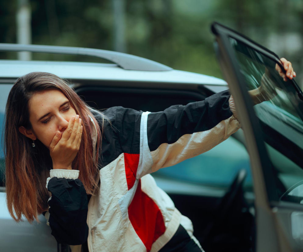Młoda kobieta z zamkniętymi oczami, oparta o otwarte drzwi samochodu, z dłonią zakrywającą usta, odczuwa mdłości podczas podróży. Dowiedz się więcej o przyczynach i łagodzeniu choroby lokomocyjnej na portalu Poradnik Zdrowie.