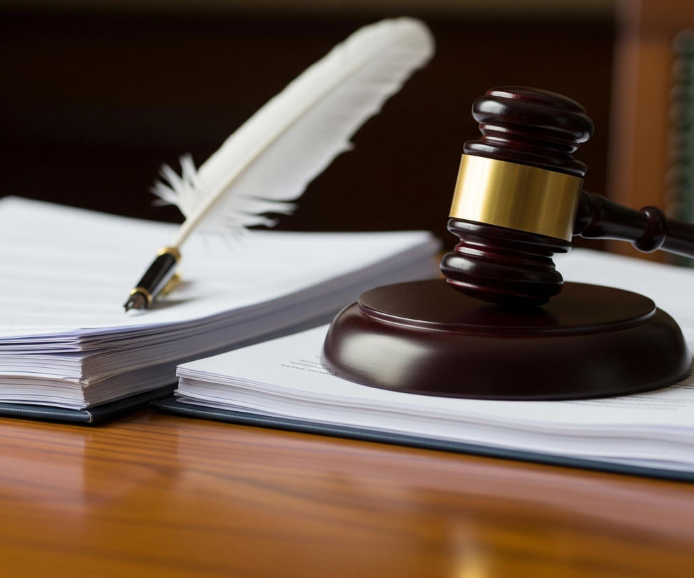 Na pierwszym planie, po prawej stronie, leży drewniany młotek sędziego w kolorze ciemnobrązowym, z poziomo umieszczoną, złotą obręczą na głowicy, oparty na okrągłej podstawce w tym samym odcieniu. Młotek spoczywa na stosie białych kartek papieru, z tekstem wydrukowanym czcionką w odcieniach szarości, z których część jest spięta w czarną okładkę. Po lewej stronie, również na stosie białych kartek, leży białe pióro, osadzone w złoto-czarnej obsadce z czarną stalówką, skierowane w stronę tekstu. Tło jest rozmyte, z widocznym brązowym meblem i fragmentem zielonego, skórzanego oparcia krzesła po prawej stronie. Całość kompozycji spoczywa na błyszczącym, drewnianym blacie w ciepłym odcieniu brązu.