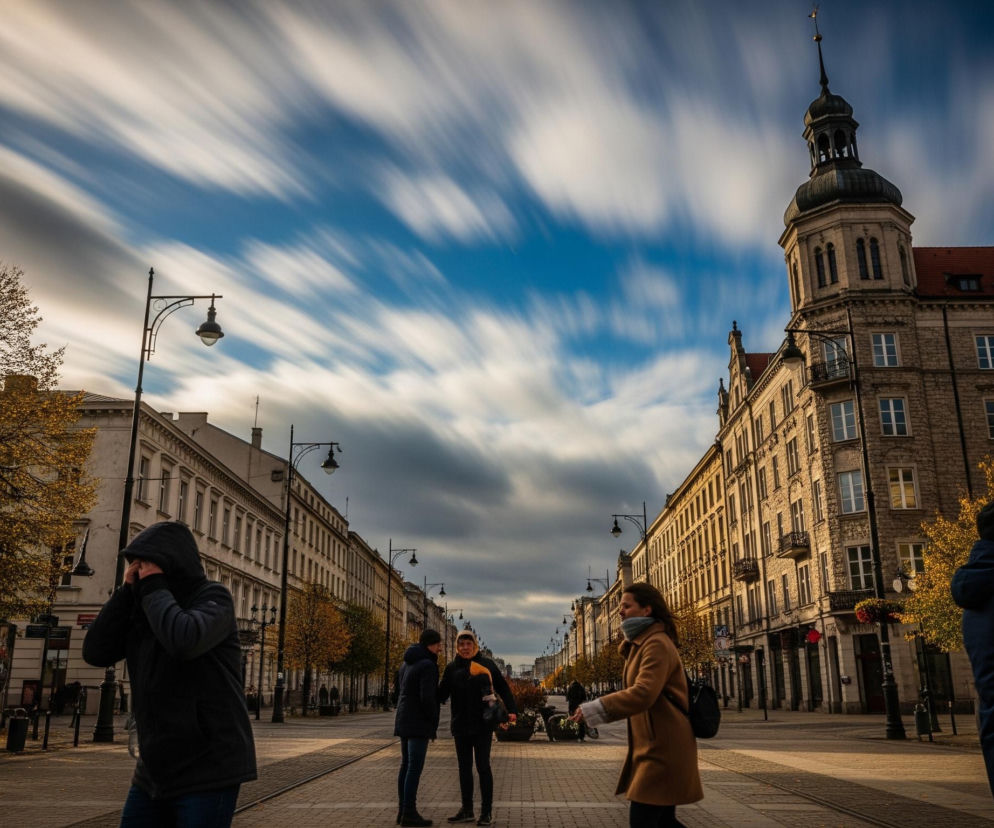 Ulica miejska rozciąga się w głąb obrazu, otoczona po obu stronach wysokimi, zabytkowymi budynkami o jasnych fasadach, z widocznymi licznymi oknami i detalami architektonicznymi. Po prawej stronie w oddali dominuje budynek z charakterystyczną, ciemną kopułą i iglicą. Na pierwszym planie, w centrum kadru, widać trzy osoby stojące w grupie, dwie z nich rozmawiają, a jedna idzie w prawo, ubrana w jasnobrązowy płaszcz i szary szalik.

Po lewej stronie znajduje się rząd drzew o liściach w jesiennych barwach, częściowo bezlistnych, a pod nimi ławki. Wzdłuż ulicy rozmieszczone są latarnie uliczne z czarnymi, ozdobnymi słupami. Niebo nad ulicą jest częściowo błękitne, z długimi, rozmytymi pasmami białych chmur, które rozciągają się horyzontalnie, tworząc efekt ruchu.
