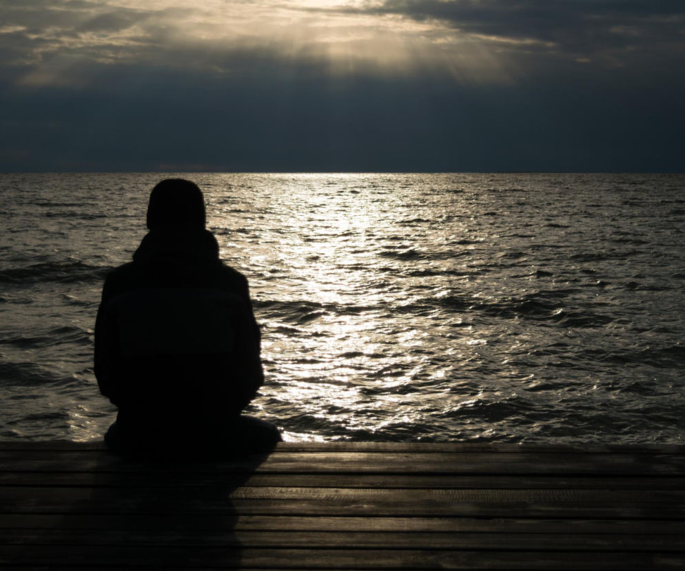Samotna sylwetka osoby siedzącej na drewnianym pomoście, tyłem do widza, wpatrującej się w morze o zmierzchu. Promienie słońca przebijają chmury, tworząc świetlistą smugę na wzburzonej wodzie. Scena symbolizuje izolację i negatywny wpływ samotności na zdrowie. Na Poradnik Zdrowie znajdziesz więcej o ryzyku zgonu i chorób związanych z brakiem więzi społecznych.