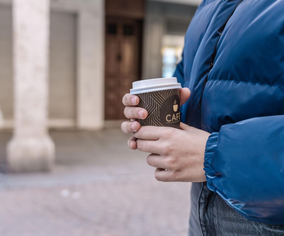 Osoba w niebieskiej kurtce trzyma w dłoni kubek kawy na wynos, co odzwierciedla poranny rytuał. Zdjęcie ilustruje problem mikroplastiku w jednorazowych kubkach, o czym można przeczytać na portalu Poradnik Zdrowie.