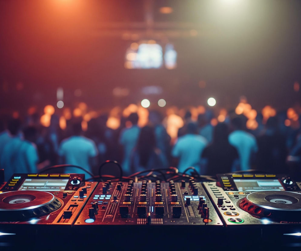 DJ-ski stół mikserski z dwoma gramofonami dominuje na pierwszym planie, oświetlony od dołu niebieskimi i zielonymi światłami. Na stole leżą słuchawki, a całe urządzenie wypełnione jest pokrętłami, suwakami i przyciskami. W tle, rozmyta i pogrążona w czerwono-pomarańczowym świetle, widoczna jest publiczność, składająca się z wielu postaci, których sylwetki są słabo zarysowane.