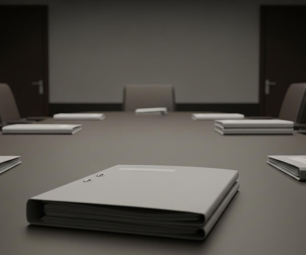 Centralnie na pierwszym planie widoczny jest pojedynczy, szary segregator leżący na długim, ciemnym stole konferencyjnym. Po obu stronach stołu ustawione są puste krzesła z ciemnymi obiciami, a na blacie leży kilka innych segregatorów, również w kolorze szarym, rozmieszczonych równomiernie. W tle, na ścianie w kolorze szarym, widoczne są dwa ciemne drzwi oraz dwie prostokątne ramy bez widocznej zawartości, całość utrzymana jest w stonowanej, szarej kolorystyce.