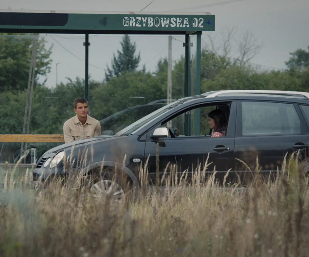Mateusz Mostowiak siedzący na ławce przystanku Grzybowa 02 w Grabinie, obok którego zatrzymuje się Lilka w samochodzie, patrząc na niego z otwartego okna. Zdjęcie ilustruje scenę z serialu M jak miłość, o której możesz przeczytać więcej na Super Seriale.