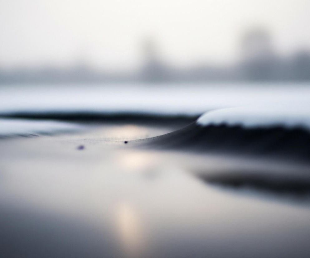 Płynna, ciemna powierzchnia odbija jasne światło, tworząc refleksy przypominające zachód słońca. Na pierwszym planie, wzdłuż lewego brzegu, widać ciemną linię, która rozszerza się w głąb obrazu. Prawa strona obrazu jest pokryta warstwą białego puchu, prawdopodobnie śniegu, który tworzy delikatny, falujący kształt. W tle, na horyzoncie, rozciąga się niewyraźny zarys ciemnych obiektów, które zlewają się z szarym, zamglonym niebem.