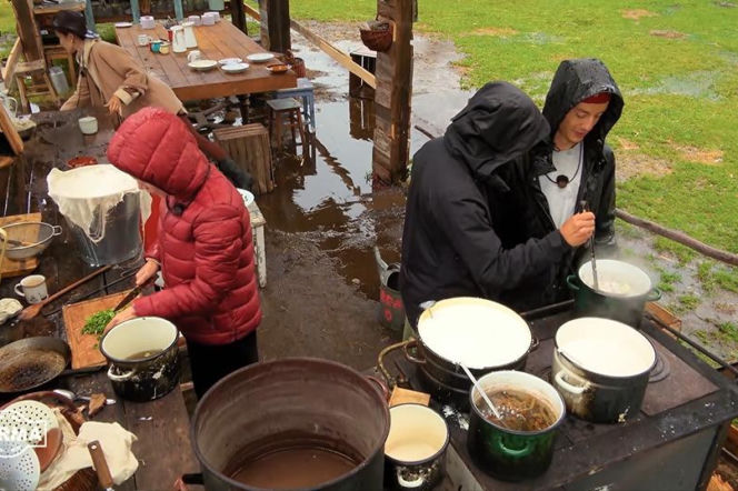 Uczestnicy programu Farma podczas przygotowywania posiłku na zewnątrz w czasie ulewy. Osoba w czerwonej kurtce kroi zioła, inna w czarnej kurtce miesza coś w garnku na piecu. Wokół widać kałuże i błoto. Scena z odcinka Farma 5, gdzie Super Seriale relacjonują trudne warunki.