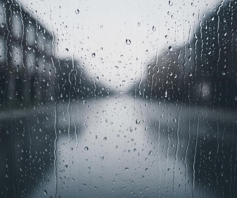 Szyba pokryta jest licznymi kroplami deszczu o różnych rozmiarach, tworzącymi smugi w dół. Za szybą widać rozmytą perspektywę ulicy z ciemnymi, niewyraźnymi budynkami po lewej i drzewami po prawej stronie. Centralna część obrazu przedstawia jasną, zamgloną przestrzeń, prawdopodobnie ulicę, która rozciąga się w oddali pod pochmurnym niebem. Całość utrzymana jest w chłodnej, szaro-niebieskiej tonacji.
