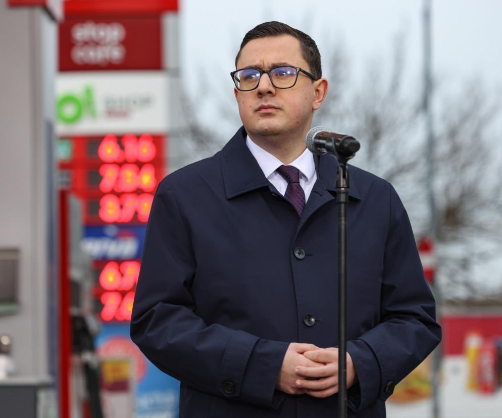 Miłosz Motyka, minister energii, w okularach i granatowym płaszczu, stoi przy mikrofonie, patrząc w prawo. W tle widać pylon stacji paliw z czerwonymi cyframi cen. Czytaj więcej o maksymalnych cenach paliw na Super Biznes.