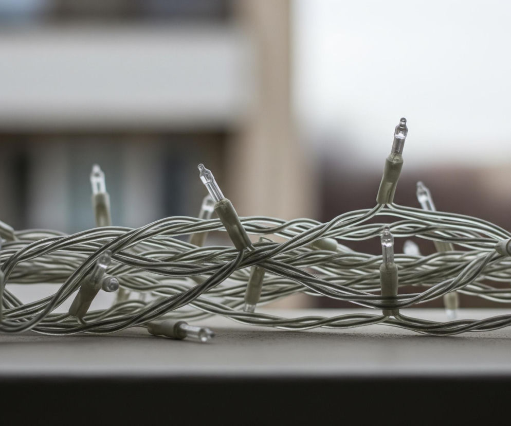 Na pierwszym planie leży zwinięty sznur drobnych lampek choinkowych o jasnoszarych przewodach. Małe, przezroczyste żarówki rozmieszczone są w regularnych odstępach wzdłuż splątanych kabli. Tło jest rozmyte, ukazując części jasnego budynku o beżowo-białej elewacji i ciemniejszych oknach, z niewyraźnym niebem lub dalekim krajobrazem u góry.